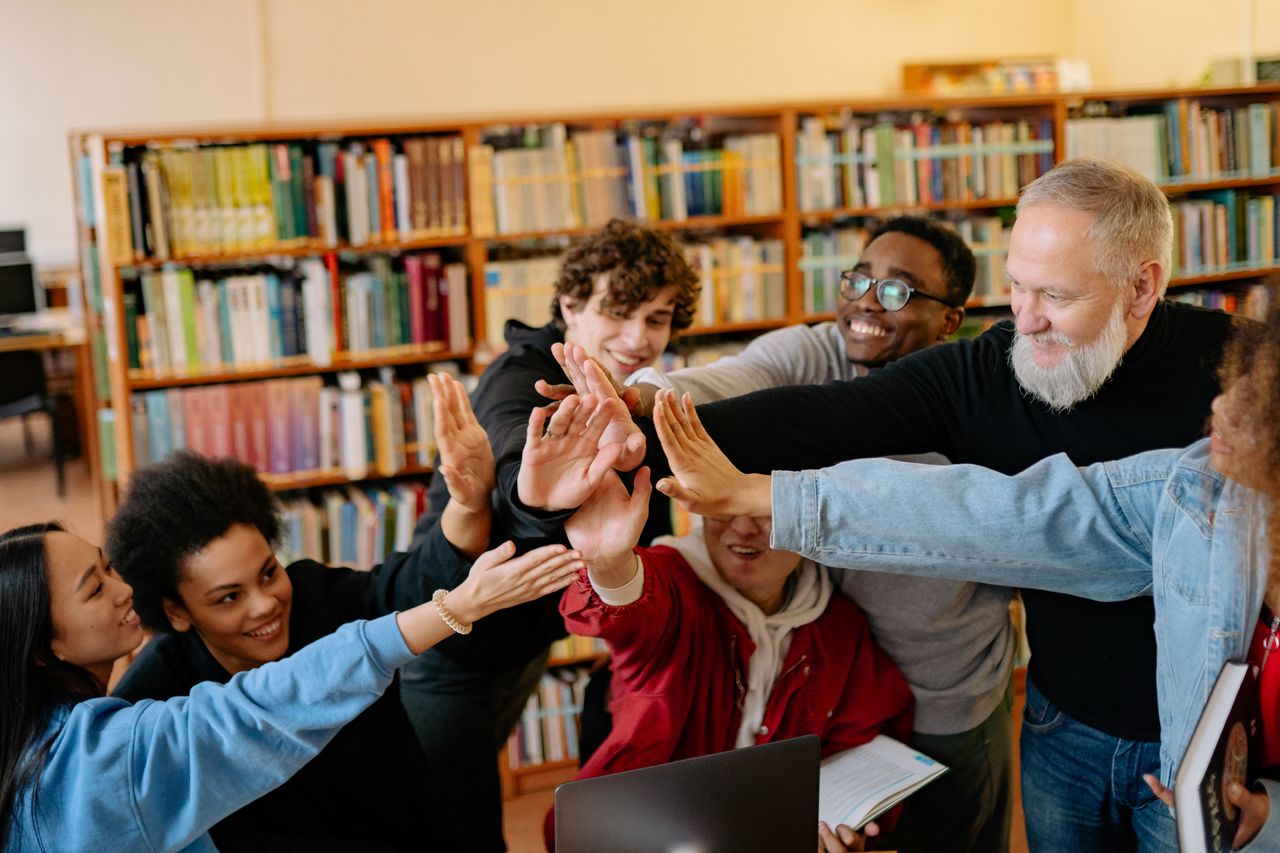 Hello world! Diverse group of students and a teacher high-fiving in a library. Celebrating teamwork and education.